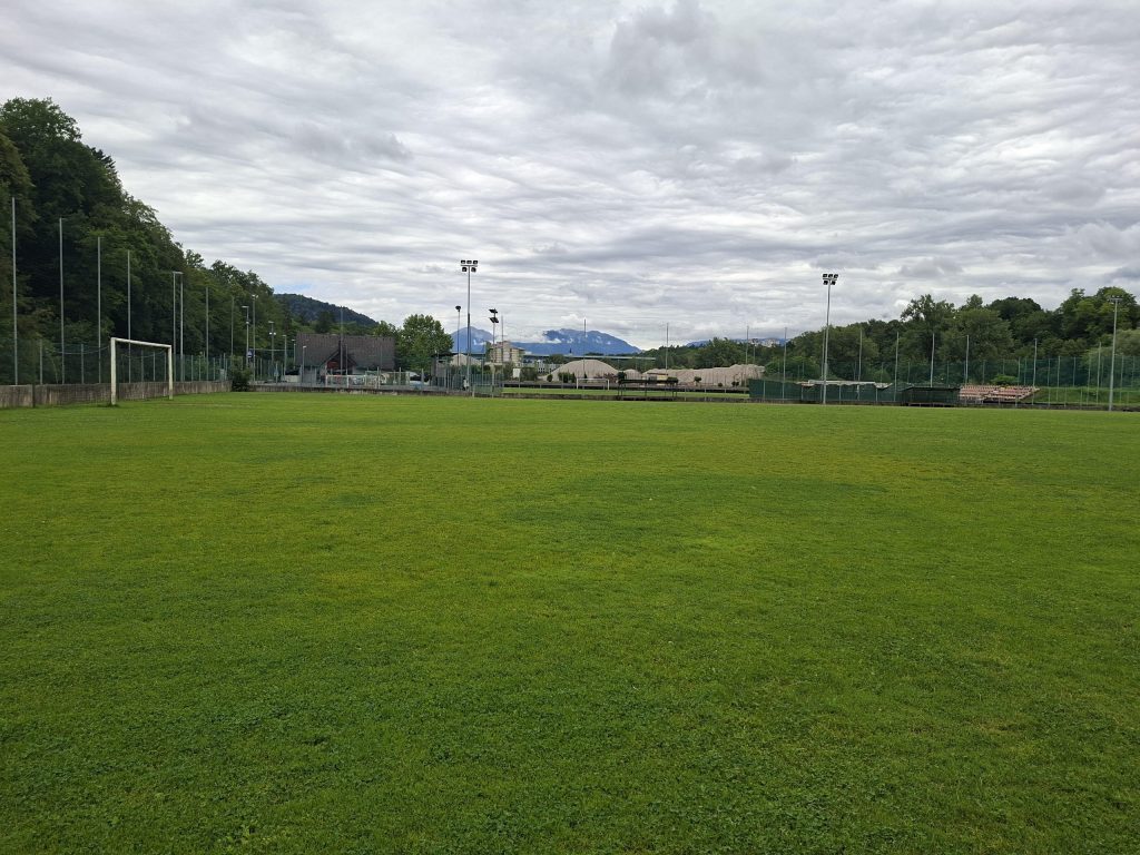 Pomožno travnato igrišče z golom, v ozadju klubski objekti in gore